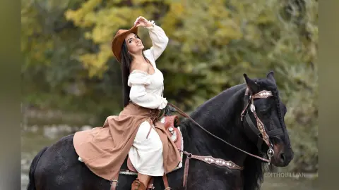 Woman in white peasant blouse and brown hat riding a black horse through a lush green forest.
