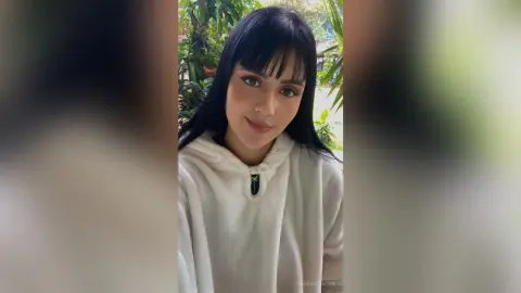 Smiling young woman with long black hair wearing a cozy white fleece hoodie in a lush garden setting.