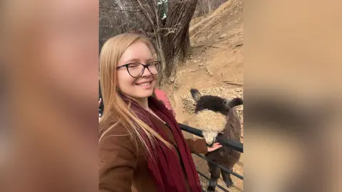 Smiling blonde woman in a brown coat and red scarf petting a black and white alpaca at an outdoor farm.