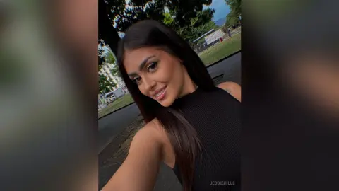 Smiling young woman in a black sleeveless top posing outdoors in a sunny park setting.