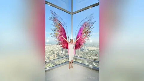 Dreamy woman in white dress with large pink angel wings standing on a high-rise balcony overlooking a city skyline.