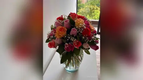 SaraBeleno displays a large, colorful bouquet of red, pink, orange, and peach roses in a clear glass vase.
