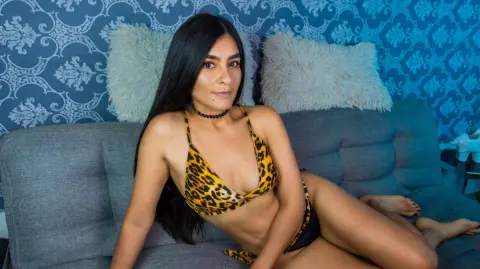 Young woman in a leopard print bikini relaxing on a gray sofa against a blue damask wallpaper background.