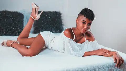 AnnPrice lies on a white bed wearing a white off-the-shoulder top and light-colored high heels, posing with one leg raised against dark pillows.