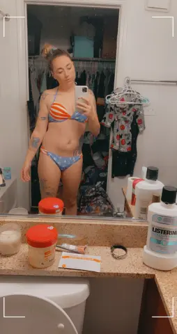 Tattooed woman in a striped bikini taking a mirror selfie in a bright bathroom.