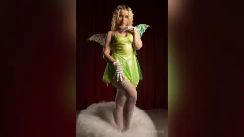 Blonde woman in a green fairy costume and wings posing on white clouds against a dark red backdrop.