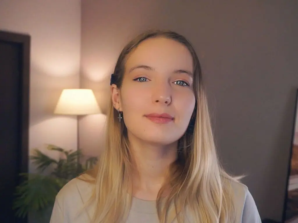 Blonde woman in a casual beige shirt smiling in a warm, dimly lit indoor setting.