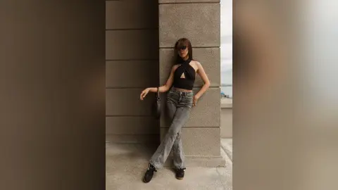 Stylish woman in a black halter top and grey jeans leaning against a concrete wall in a minimalist urban setting.