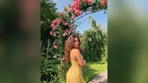 InesMoore poses in a yellow floral dress, standing under a pink rose arch in a lush green garden.