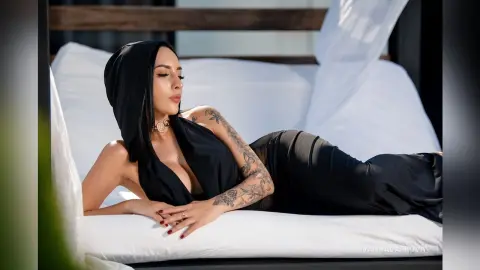 Elegant woman in a black halter dress relaxing on a white outdoor lounge bed in bright sunlight.