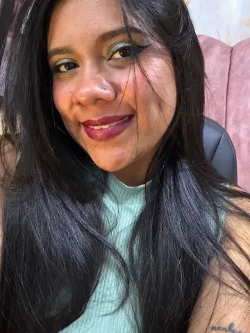 Smiling woman with long dark hair and green eyeshadow wearing a light mint green ribbed top in a cozy setting.