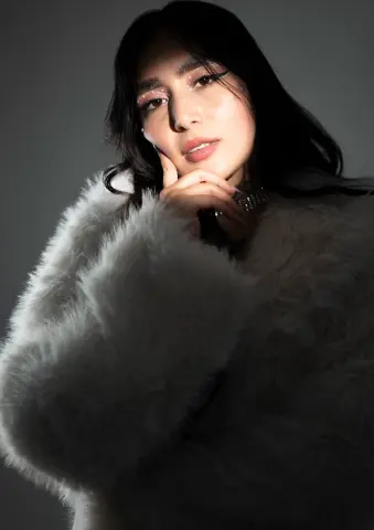 Young woman with glamorous makeup wearing a cozy white faux fur coat in a moody studio setting.