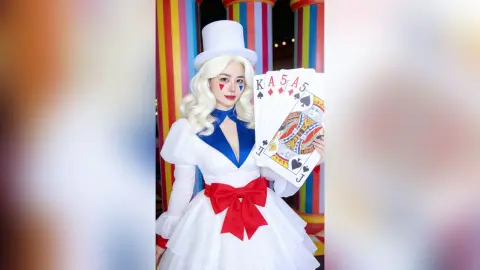 Blonde woman in a white Alice in Wonderland queen costume and top hat holding playing cards in a colorful setting.