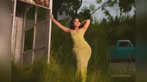Elegant woman in a long olive green dress posing in a grassy field near a vintage white door at sunset.