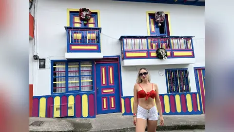 Blonde woman in a red crop top and white shorts standing before a colorful blue and yellow Mediterranean building.