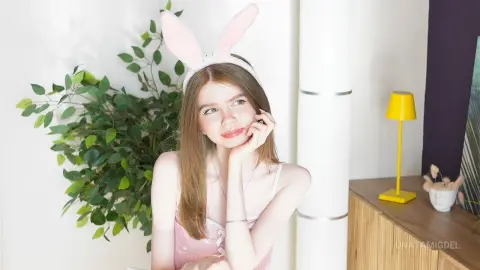 Young woman with bunny ears in a pink outfit smiling in a bright, cozy room with green plants.