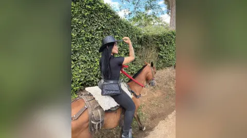 Stylish woman in black equestrian outfit and cowboy hat riding a brown horse through a lush green garden.