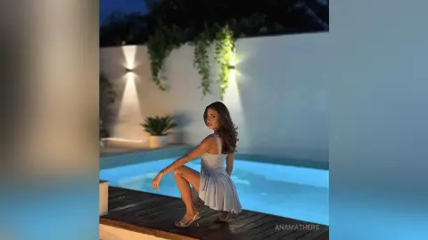 Elegant woman in a white summer dress sitting by a luxury swimming pool at night under soft garden lighting.