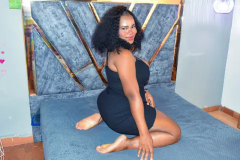 SkyllerFoxy — Smiling woman in a black dress sitting on a grey velvet bed in a modern bedroom setting.