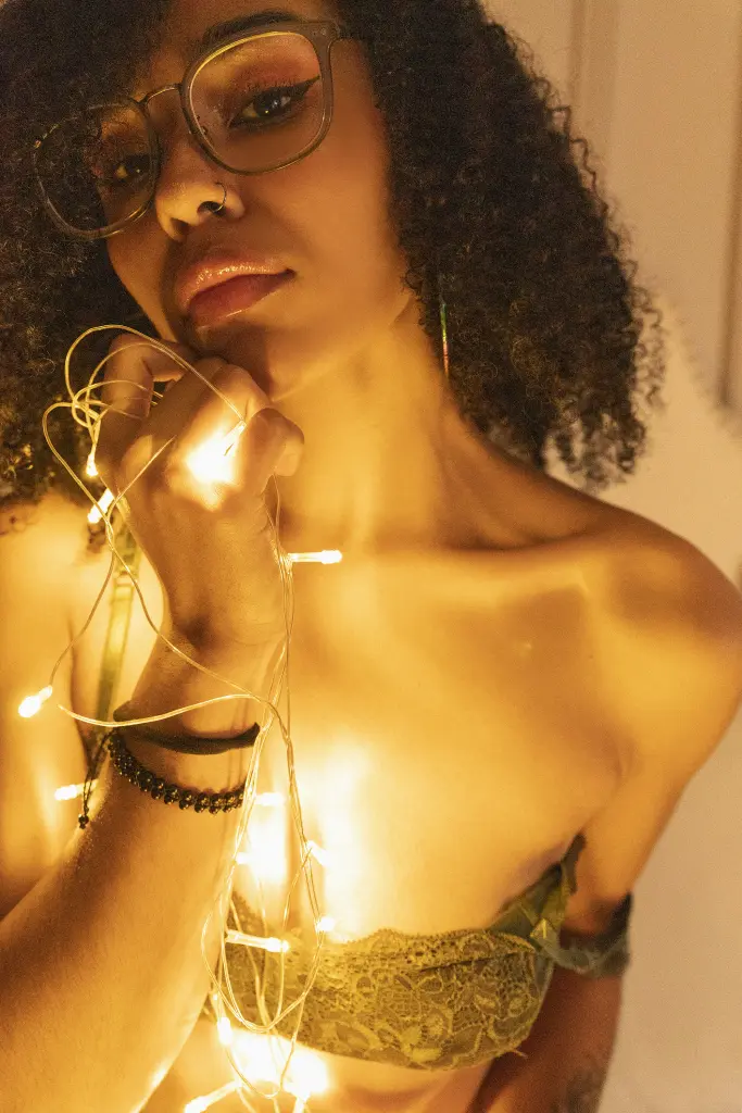 MacarenaaWalker — Young woman with curly hair and glasses wearing lace lingerie, illuminated by warm fairy lights in a cozy mood.