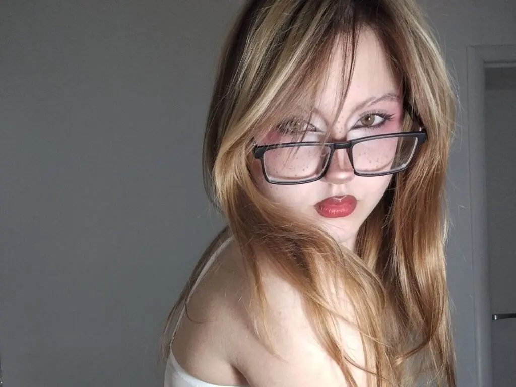 Blonde woman with glasses and red lipstick posing in a minimalist indoor setting with a soft aesthetic mood.