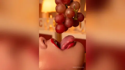 Close-up of a woman with red lipstick licking a fresh bunch of purple grapes in warm, moody lighting.
