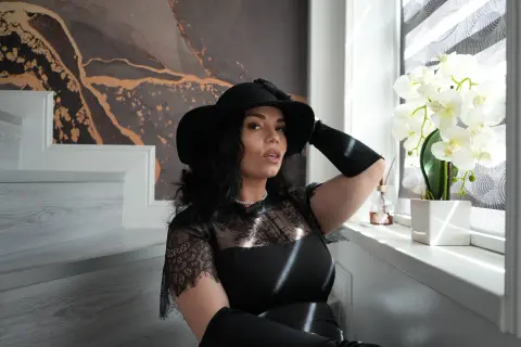 Elegant woman in a black lace dress and wide-brimmed hat sitting by a window with white orchids.