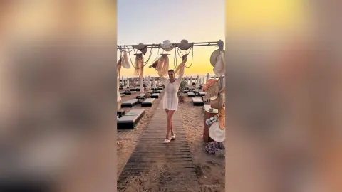 Woman in a white summer dress standing on a wooden boardwalk at a beach club during sunset.
