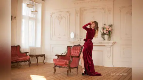 Elegant woman in a long red velvet gown posing in a luxurious white classical room with vintage furniture.