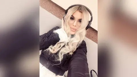 Blonde woman in a black puffer jacket and white sweater wearing headphones in a modern indoor setting.