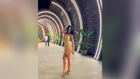 NicoleBella stands in a curved, lit hallway wearing a gold cut-out bodysuit and gold high heels.