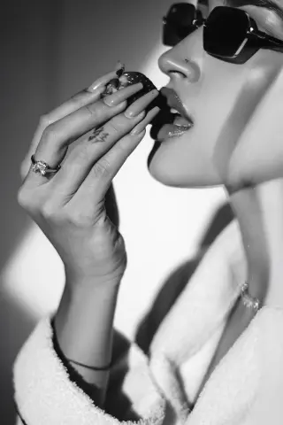 Stylish woman in a white robe and sunglasses eating a dark fruit in a bright, moody black and white portrait.
