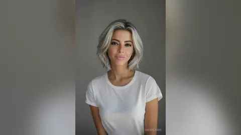 Smiling woman with wavy silver hair wearing a casual white t-shirt against a neutral gray background.