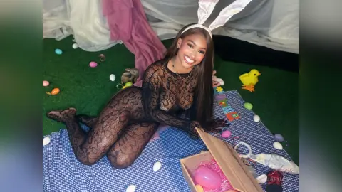 LiaTyler smiles while wearing a black lace bodysuit and bunny ears, posing on a blue blanket surrounded by Easter eggs and a small yellow chick.