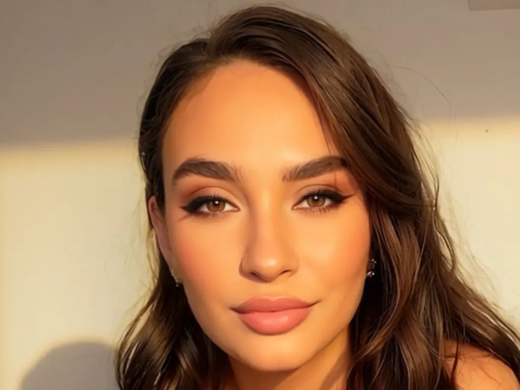 Close-up portrait of a young woman with warm makeup and wavy brown hair in soft sunlight.