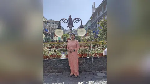 ManishaJi — ManishaJi stands outdoors in a peach-colored long dress next to a large decorative street lamp.
