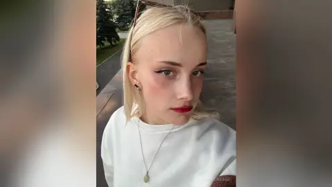 Blonde woman in a white sweatshirt and silver necklace posing outdoors with a soft, natural look.