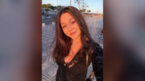 Smiling young woman in a black long-sleeve top enjoying a warm sunset outdoors.