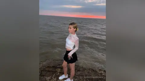 Blonde woman in a white lace top and black skirt standing by the ocean during a peaceful sunset.
