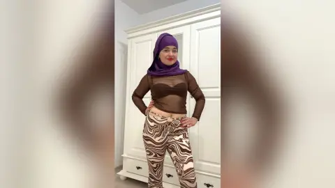 Confident woman in a purple hijab, brown sheer top, and patterned trousers posing in a bright room.