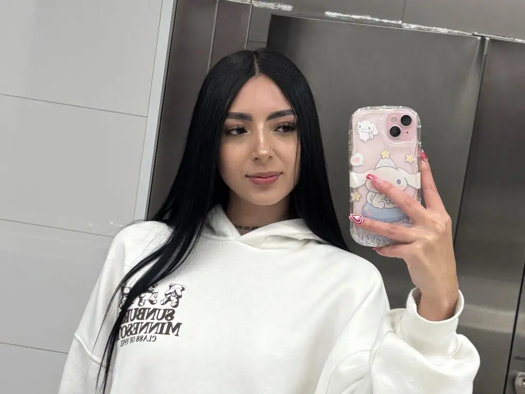 Young woman with long dark hair wearing a white graphic hoodie taking a mirror selfie in a modern room.