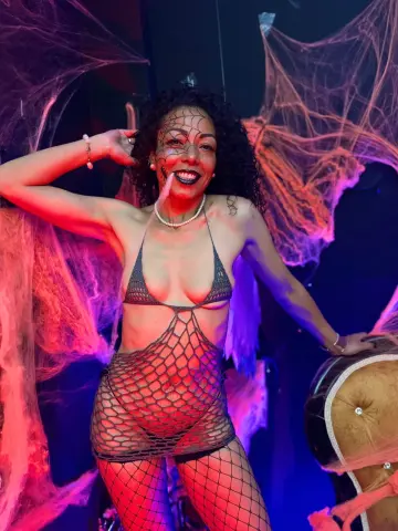 Smiling woman in black fishnet lingerie posing amidst spooky spiderwebs in moody purple lighting.