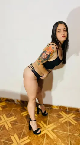 Tattooed woman in black lingerie posing against a white wall on a patterned tile floor.