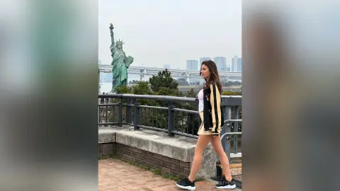 AnaMathers stands on a balcony overlooking a city skyline, wearing a white top and a black and gold striped jacket with a tan skirt.