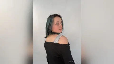 SoleneWinter looks over her shoulder with her blue-tinted hair, wearing a black off-the-shoulder top against a plain white background.