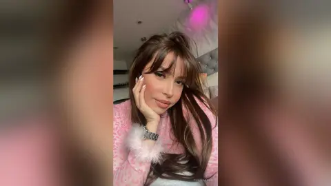 Young woman with long brown hair wearing a pink leopard print sweater in a cozy, soft-lit bedroom setting.