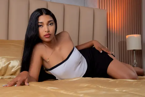 JasminMale lies on a gold bed wearing a white tube top and black shorts, posing with her long black hair down.