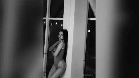 MilanaVolkova poses in a tight bodysuit against a white pillar, looking back over her shoulder with long dark hair.