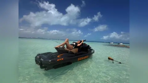 Woman in black swimwear relaxing on a jet ski in turquoise tropical ocean water under a blue sky.