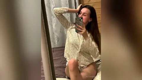Young woman in white floral loungewear taking a mirror selfie in a cozy bedroom setting.